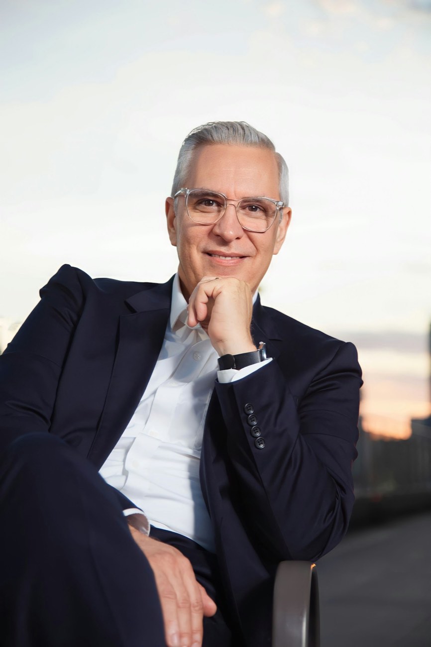 A grey-haired man in a dark suit and glasses, seated with his hand to his chin