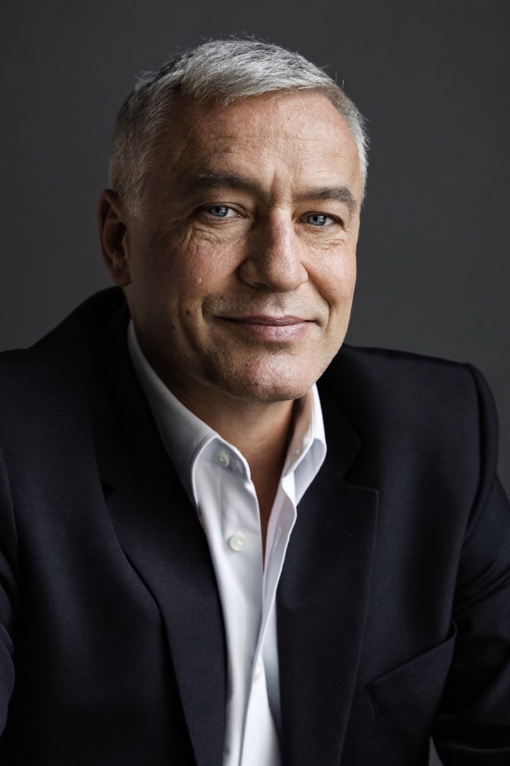 A silver-haired man in a navy suit and open white shirt, looking into the camera