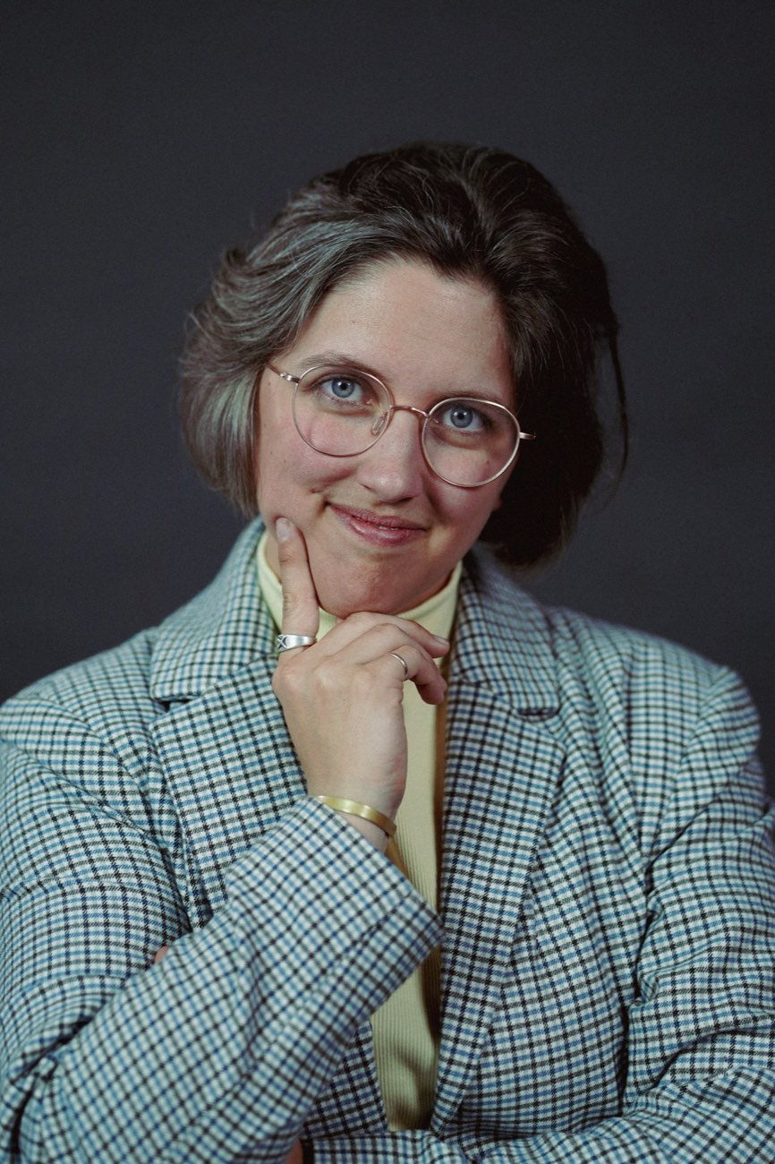 A woman in a checked blazer and glasses, her hand to her chin