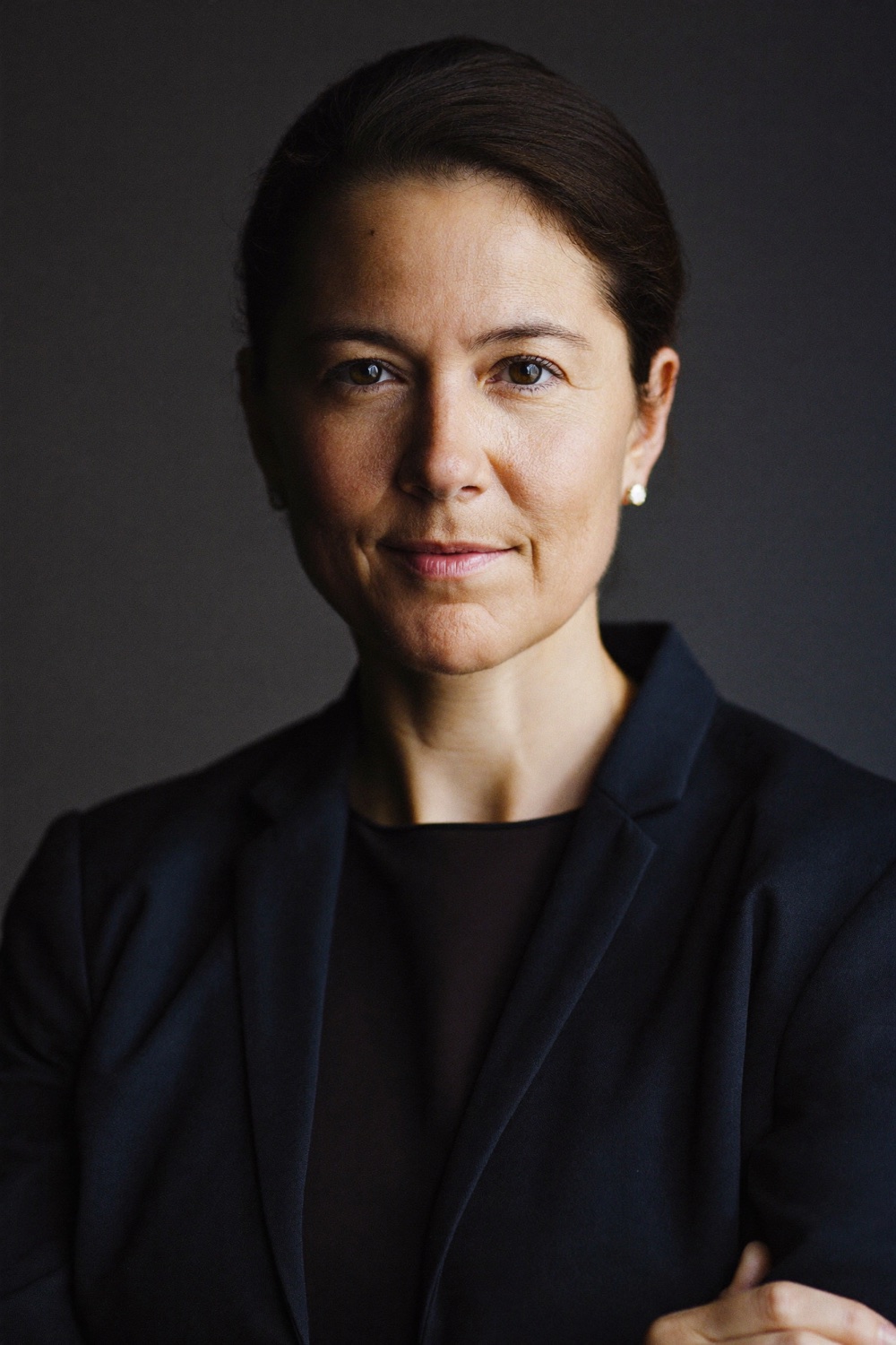 A woman with dark hair tied back, in a navy blazer, arms folded