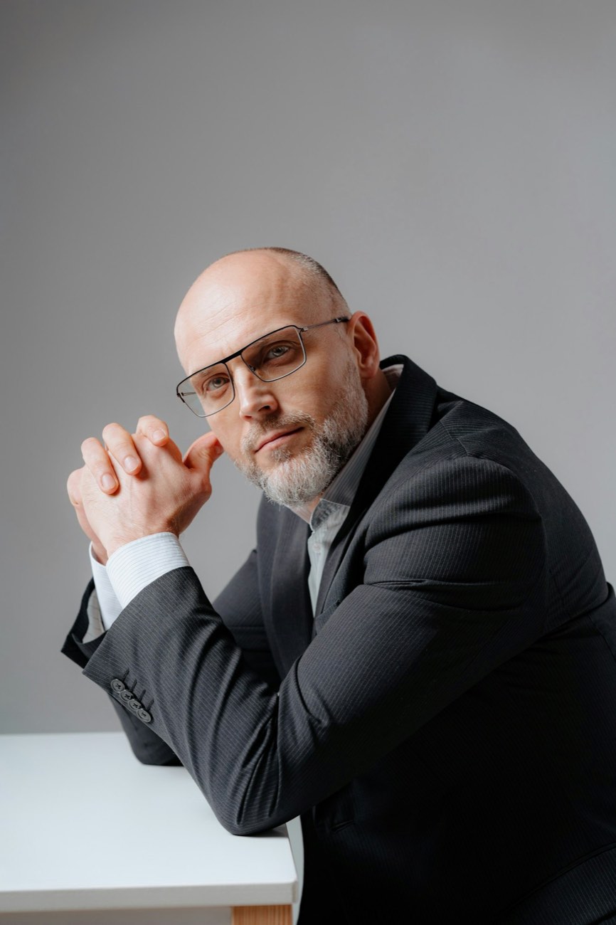 A man in a dark blazer and glasses, hands clasped at a desk